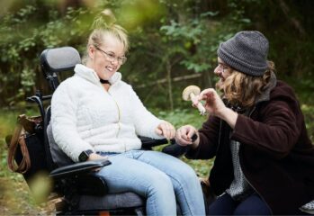 Ung kvinne i rullestol sammen med BPA-assistent i skogen.