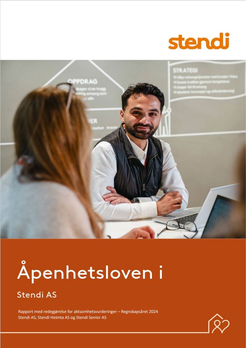 To ansatte sitter rundt et møtebord og jobber. Den ene ser i kameraet og smiler.