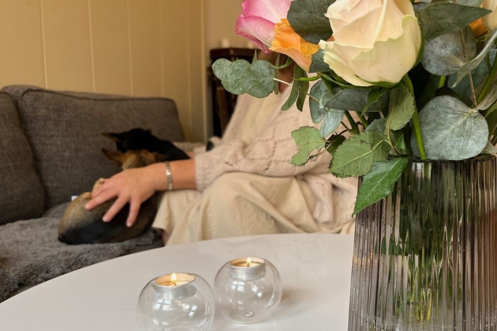 Blomster og levende lys på et bord. Enslig fostermor koser med hundene sine i bakgrunnen.