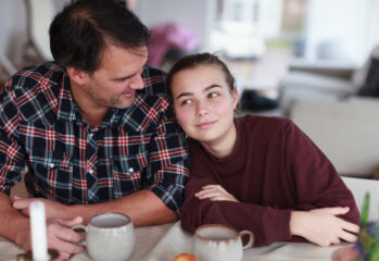 Tenåringsjente lener seg mot skulderen til sin fosterfar ved kjøkkenbordet