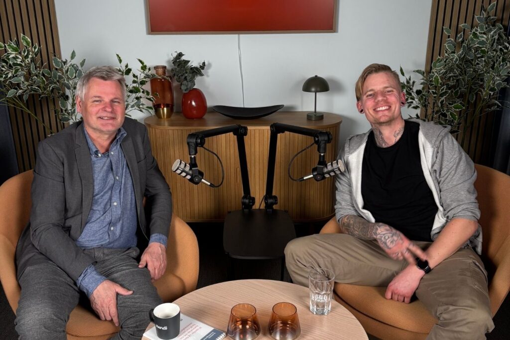 To menn i studio. En med grått hår og dress. Den andre med hettegenser og tatoveringer.